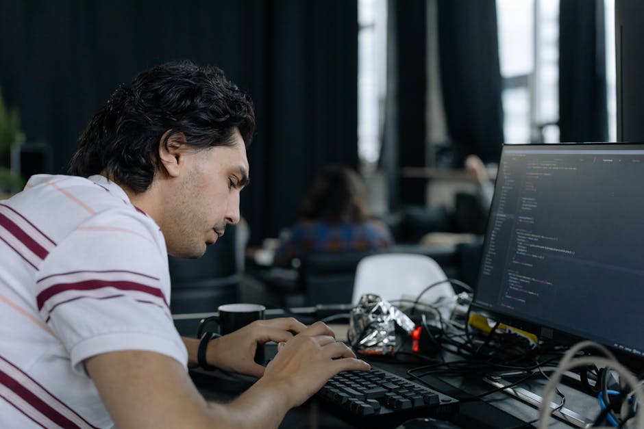 A young man with dark, wavy hair sits at a desk in a modern office space, focused on coding on his computer monitor displaying lines of programming text. The desk is cluttered with tangled wires, a cr
