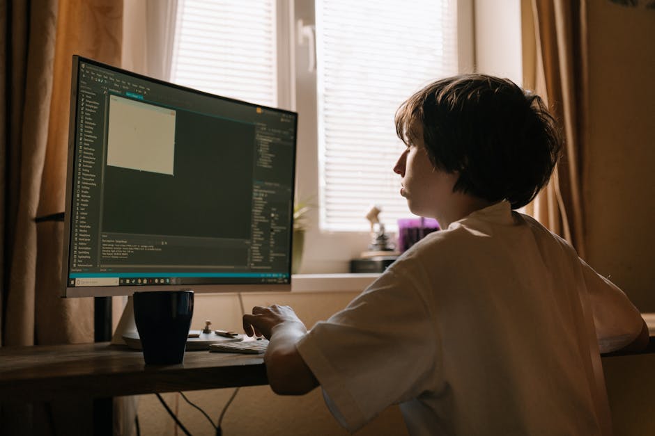 A young man with dark, wavy hair sits at a desk in a modern office space, focused on coding on his computer monitor displaying lines of programming text. The desk is cluttered with tangled wires, a cr