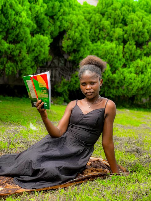 A young woman with dark skin and natural hair styled in a puff, sitting on a patterned blanket on the grass in a garden or park setting. She is wearing a black sleeveless dress with thin straps and is